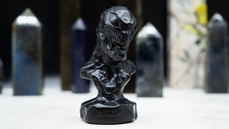 May include: A black bust sculpture of a character with a detailed face and chest. The sculpture is set against a blurred background of various crystal towers. The bust has a textured surface and is made of a dark, possibly obsidian, material.