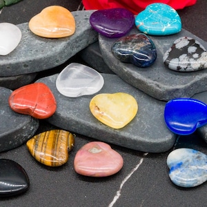 Könnte beinhalten: Eine Sammlung herzförmiger Edelsteine in verschiedenen Farben, darunter Weiß, Orange, Lila, Türkis, Schwarz und Gelb. Die Steine sind auf grauen, strukturierten, rechteckigen Plattformen angeordnet. Die Edelsteine variieren in Größe und Textur.