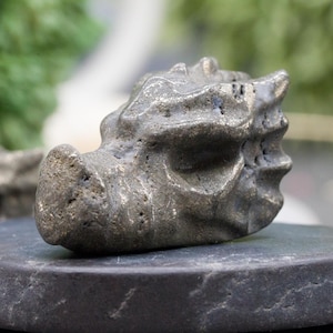 May include: Three small, gray, stone-like sculptures with a rough, textured surface. The sculptures are arranged on a black, round stone base.
