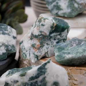 May include: A collection of moss agate stones in various shapes and sizes. The stones are a mix of white, green, and brown colors. The stones are arranged on a wooden surface.