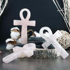 May include: Rose quartz ankhs in various sizes are displayed on a wooden surface. The ankhs, a symbol of life, are a soft pink color and have a polished, smooth texture. The background includes cotton bolls and dark accents.