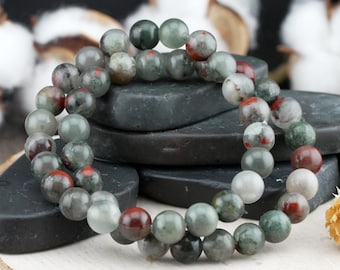 May include: A beaded bracelet composed of smooth, spherical stones in shades of sage green, dark forest green, and translucent grey, accented with distinct spots of deep reddish-brown. The beads are strung together in a circular, elasticized band.