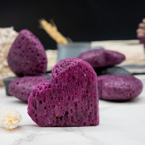May include: Purple heart-shaped bath sponges, made from natural materials, are arranged on a white surface.