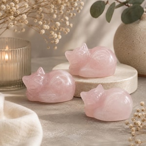 May include: Three pink rose quartz cat figurines in a sleeping pose. The cats are smooth and polished, with visible details. The figurines are displayed on a neutral surface, with a candle and vase in the background.