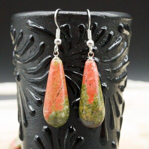 May include: A pair of teardrop-shaped earrings featuring polished unakite stone with mottled patterns of salmon pink and olive green. The stones are suspended from silver-toned metal fishhook ear wires, creating a dangling design.