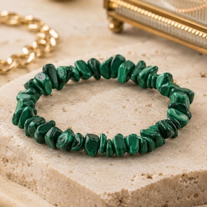 Puede incluir: Un brazalete de virutas de malaquita. El brazalete está hecho de piedras de malaquita pulidas, de forma irregular y de color verde oscuro. Se muestra sobre una superficie clara y texturizada.
