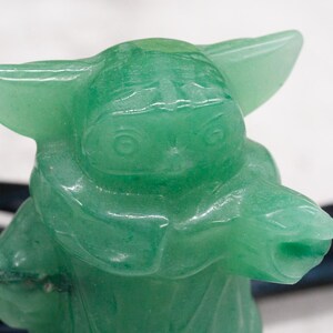 以下が含まれることがあります： 大きな耳とローブを着たキャラクターの緑色の置物。置物は半透明の明るい緑色の素材でできています。フィギュアは、黒のアクセントが付いた白い背景に配置されています。