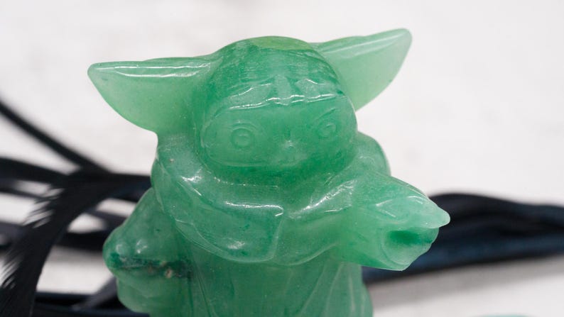 以下が含まれることがあります： 大きな耳とローブを着たキャラクターの緑色の置物。置物は半透明の明るい緑色の素材でできています。フィギュアは、黒のアクセントが付いた白い背景に配置されています。