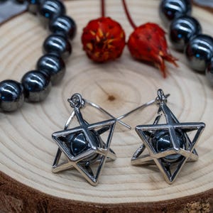 May include: Silver-coloured star earrings with a dark grey sphere at the centre. The earrings are displayed on a wooden surface, with a beaded bracelet and dried red flowers.