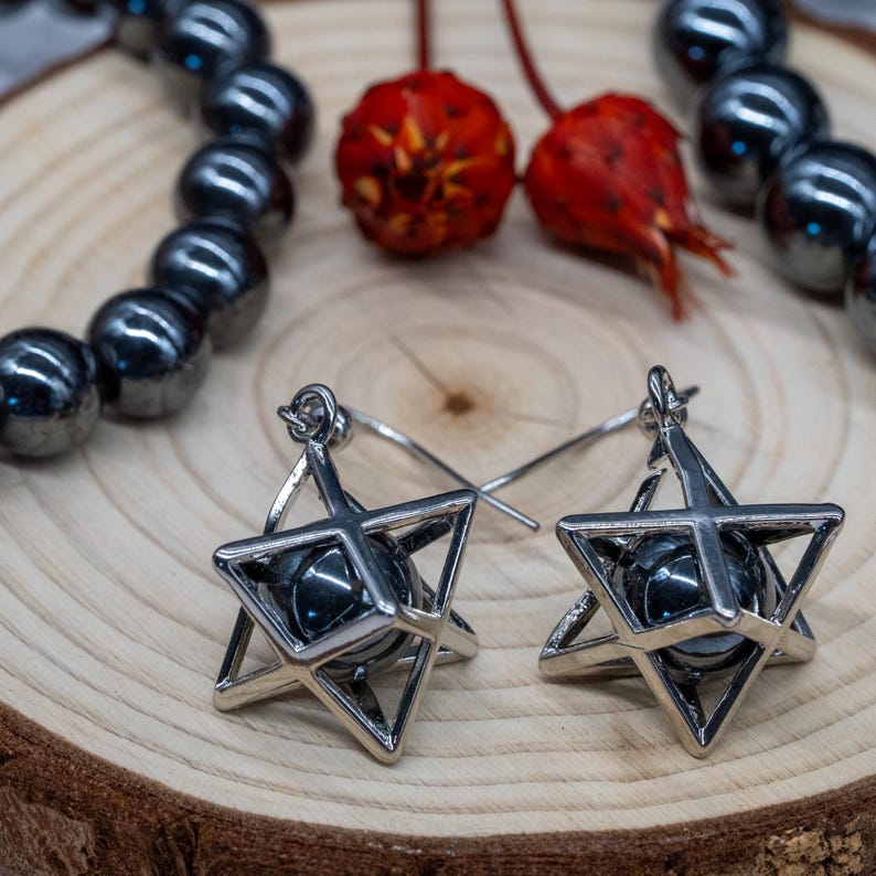 May include: Silver-coloured star earrings with a dark grey sphere at the centre. The earrings are displayed on a wooden surface, with a beaded bracelet and dried red flowers.