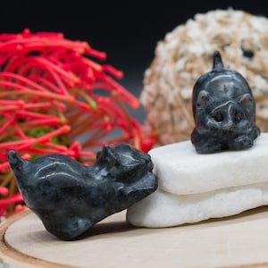 May include: Two small, dark gray cat figurines with a glossy finish. One cat is lying down, the other is upright. The figurines are on white stones, with red floral accents in the background. The cats are detailed with carved features.