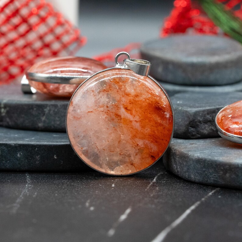 Op de afbeelding: Een close-up van een ronde hanger met een zilverkleurig metalen frame. De hanger heeft een doorschijnende, rood-oranje steen met interne adering. De hanger wordt getoond op een donker, getextureerd oppervlak.