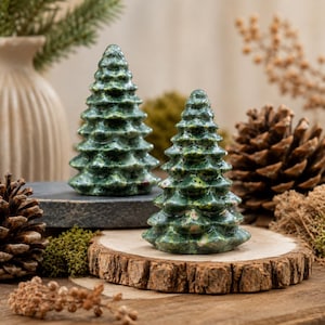 May include: Two conical tree figurines crafted from polished green stone with mottled textures. The taller tree stands on a dark gray circular base, while the shorter tree sits on a light-colored natural wood slice with visible bark edges.