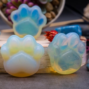 May include: Three opalescent paw-shaped stones with a translucent appearance. The stones have a mix of blue, yellow, and white hues. The paw prints are smooth and rounded, with a glossy finish. The background includes a blurred arrangement of plants.