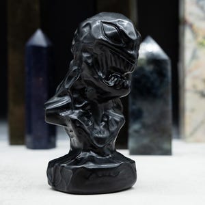 May include: A black sculpture of a character's bust with a detailed, textured surface. The sculpture is in front of several crystal obelisks of various colors and patterns, set against a dark background.