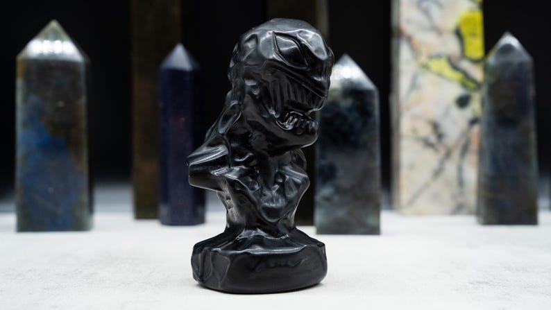 May include: A black sculpture of a character's bust with a detailed, textured surface. The sculpture is in front of several crystal obelisks of various colors and patterns, set against a dark background.