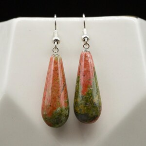 May include: A pair of drop earrings featuring polished, teardrop-shaped stones with a mottled pattern of salmon pink and olive green. The stones are suspended from silver-toned metal fishhook wires, creating a dangling design that catches the light.