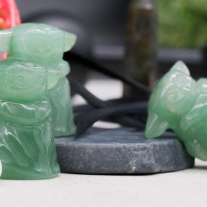 May include: Green aventurine crystal figurines of a character with large ears. One figurine is upright, and another is lying down. The figurines are on a gray stone, with a white stone and red flowers in the background.