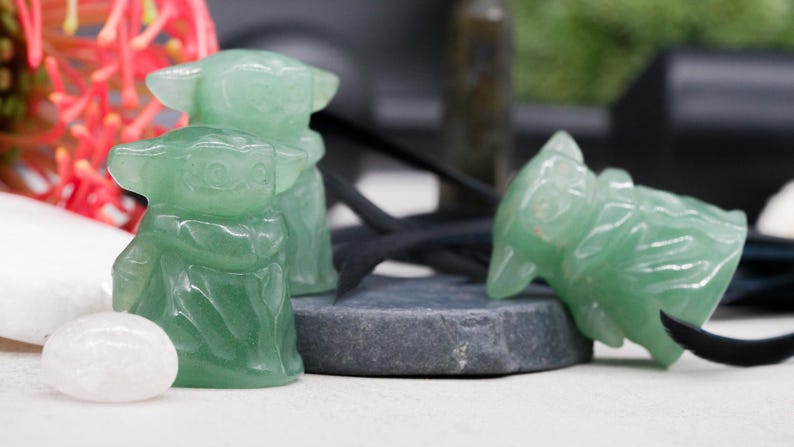 May include: Green aventurine crystal figurines of a character with large ears. One figurine is upright, and another is lying down. The figurines are on a gray stone, with a white stone and red flowers in the background.