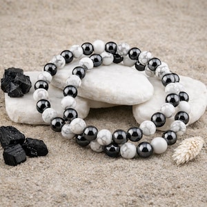 May include: Two beaded bracelets with black and white beads are displayed on a sandy surface. The bracelets are resting on white stones, with black stones and a dried plant nearby. The beads appear to be approximately 0.3 inches in diameter.