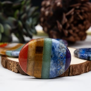 May include: A polished, oval-shaped chakra stone with a rainbow color scheme. The stone is divided into vertical stripes of red, orange, yellow, green, blue, indigo, and violet. It rests on a small wooden slice, with other stones and natural elements in the background.
