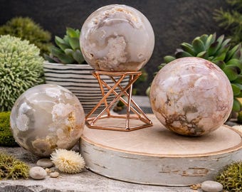May include: Three decorative agate spheres in shades of white, beige, and pink. One sphere rests on a copper-colored metal stand, while the others sit on a wooden slice and a stone surface. The spheres have unique patterns and textures.