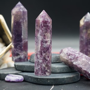May include: Several amethyst crystal towers of varying heights are displayed. The crystals are a deep purple color with white veining. They are arranged on a dark surface with small, gray stones and a decorative container in the background.