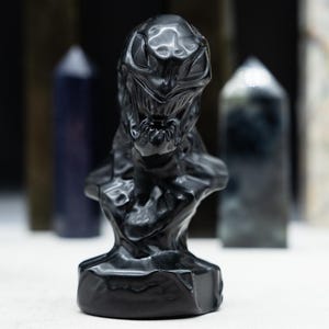 May include: A black sculpture of a creature's bust with a detailed, textured surface. The sculpture is the focal point, with several blurred, elongated crystal formations in the background. The bust has a menacing expression.