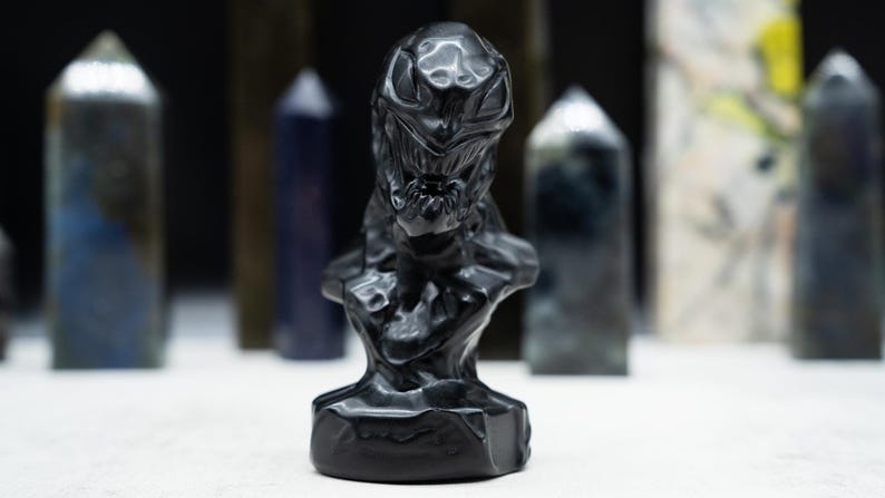 May include: A black sculpture of a creature's bust with a detailed, textured surface. The sculpture is the focal point, with several blurred, elongated crystal formations in the background. The bust has a menacing expression.