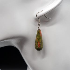 May include: A teardrop-shaped earring featuring a polished, mottled stone with a blend of olive green, forest green, and vibrant orange-red speckles. The stone is suspended from a simple silver-toned metal hook wire, creating a dangling design.