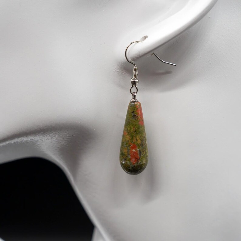 May include: A teardrop-shaped earring featuring a polished, mottled stone with a blend of olive green, forest green, and vibrant orange-red speckles. The stone is suspended from a simple silver-toned metal hook wire, creating a dangling design.