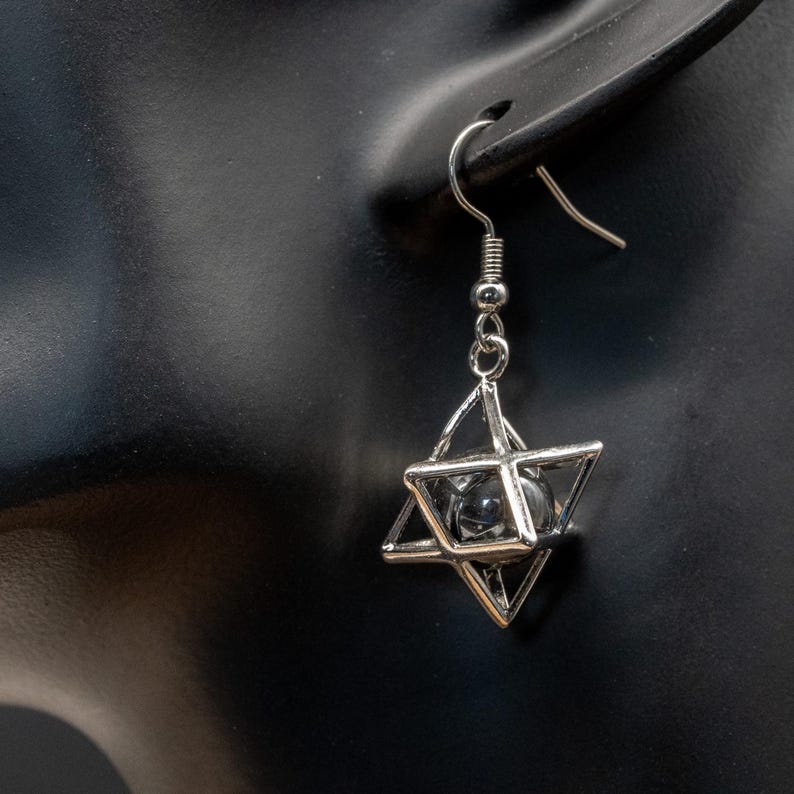 May include: A silver earring with a geometric star design, enclosing a clear, spherical bead. The earring, with a hook closure, is shown against a black backdrop. The design is intricate.
