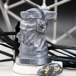 May include: A translucent gray figurine of a character from a popular science fiction franchise. The figure is standing on a white stone, with black feathers and a geometric metal structure in the background. The figurine is approximately 3 inches tall.