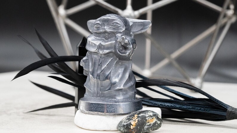 May include: A translucent gray figurine of a character from a popular science fiction franchise. The figure is standing on a white stone, with black feathers and a geometric metal structure in the background. The figurine is approximately 3 inches tall.