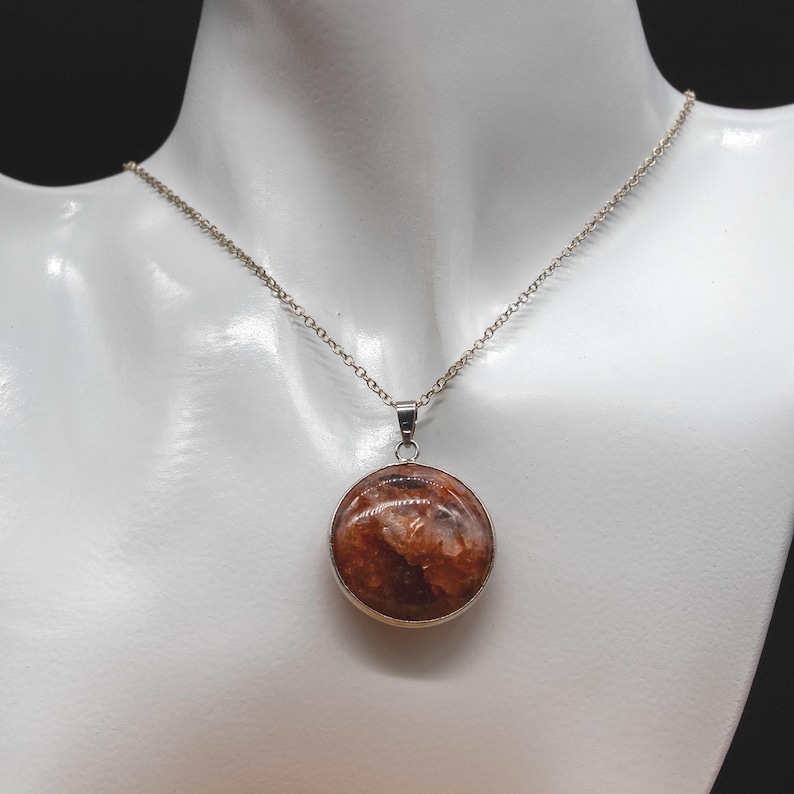 Op de afbeelding: Een zilveren ketting met een ronde hanger. De hanger heeft een roodbruine steen met lichtere insluitsels, gezet in een zilveren rand. De ketting wordt getoond op een witte mannequin.