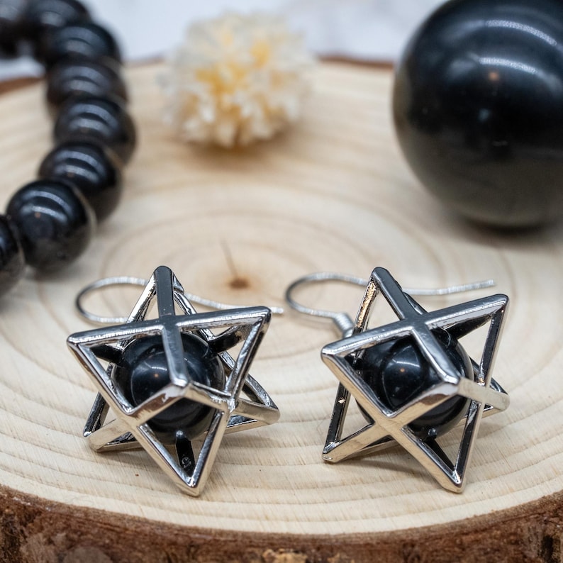 May include: A pair of silver-tone earrings featuring a star-shaped cage design with a black sphere at the center. The earrings are displayed on a wooden surface, alongside a black beaded bracelet and a black sphere.