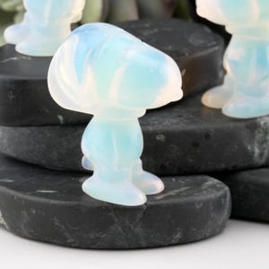 以下が含まれることがあります： 暗く質感のある表面に立っている、漫画のキャラクターの半透明でオパールのような置物。 このフィギュアは丸い頭と体をしており、足が見えます。 この素材は、青と白のヒントがあり、柔らかく光る品質を持っているようです。