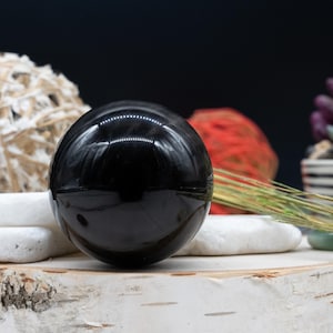 Könnte beinhalten: Eine polierte schwarze Obsidiankugel, auch bekannt als Kristallkugel, liegt auf einer weißen Holzoberfläche. Die Kugel reflektiert Licht und hat eine glatte, glänzende Oberfläche.