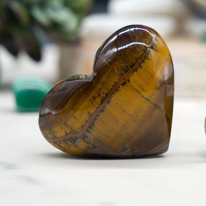 May include: A polished tiger's eye gemstone carved into a heart shape. The stone features bands of golden brown and dark brown colors. The heart is sitting on a white surface.