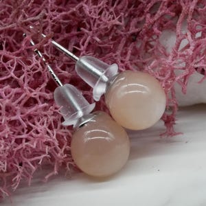Pode incluir: Um par de brincos de botão redondos cor de pêssego com pinos de prata e costas transparentes em forma de flor. Os brincos são exibidos em um fundo de material rosa texturizado.