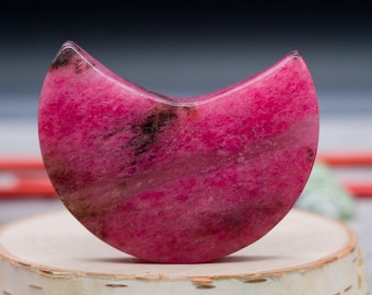 Puede incluir: Una talla en forma de media luna hecha de piedra de rodonita pulida. El objeto presenta un color base magenta y rosa vibrante con distintas inclusiones minerales irregulares de color marrón oscuro a negro en su superficie lisa y curva.