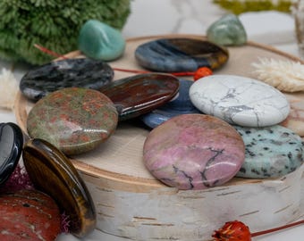 May include: A collection of polished, oval-shaped gemstones in various colors, including green, red, pink, and blue. The stones are arranged on a small, round wooden tray, showcasing their unique patterns and textures. The gemstones are a mix of sizes and shapes.