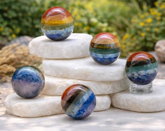May include: A collection of colorful, striped gemstone spheres in various sizes. The spheres are arranged on white, irregularly shaped stones. The colors include red, orange, yellow, green, blue, and purple. The background features green foliage and yellow flowers.