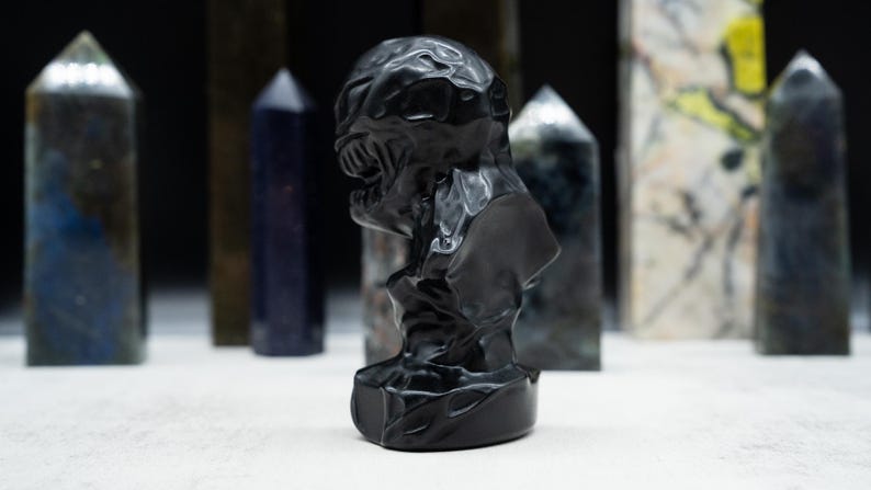 May include: A black sculpture of a creature's head and shoulders, with a detailed, textured surface. The sculpture is on a white surface, with several gemstone obelisks in the background.