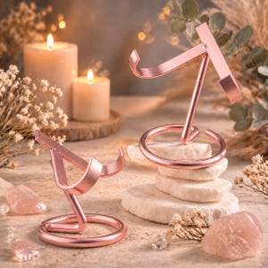 May include: Two rose gold metal phone stands. Each stand has a circular base and a curved top. The stands are on a light-coloured surface with candles and decorative elements in the background. The stands are a decorative item.