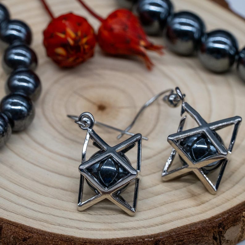 May include: Silver-coloured earrings with a geometric design. Each earring has a dark, round stone set within a star-shaped frame. The earrings are displayed on a wooden surface, with a strand of dark beads and red berries in the background.