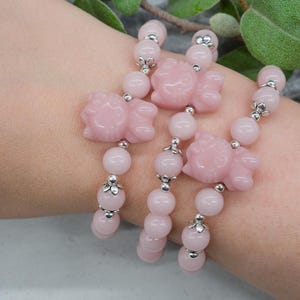 Puede incluir: Tres pulseras rosas con cuentas con dijes en forma de flor y detalles plateados. Las pulseras están hechas de cuentas redondas y se usan en una muñeca. El fondo incluye hojas verdes.