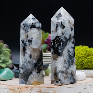 Könnte beinhalten: Zwei schwarze und weiße Kristalltürme, auch bekannt als Mondsteintürme, auf einer Holzoberfläche. Die Türme sind hoch und schlank, mit einer spitzen Spitze. Die Kristalle sind eine Mischung aus Weiß und Schwarz, mit einem schimmernden Effekt.