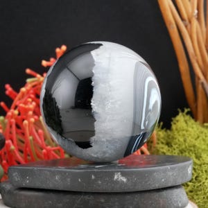 May include: A banded agate sphere, predominantly black and white, with a polished finish. The gemstone ball rests on a dark grey, layered platform. The sphere's surface reflects light, showcasing its smooth texture and natural patterns.