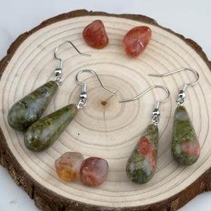 May include: Two pairs of drop earrings featuring polished, teardrop-shaped green and reddish-brown stone beads suspended from silver-toned metal hooks. Four loose, smooth, rounded reddish-brown stones are arranged around the earrings.